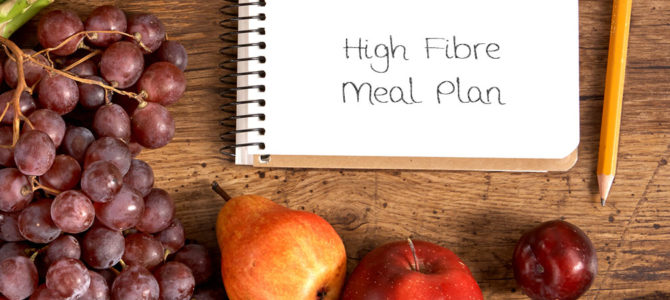 A wooden countertop with a selection of fruits and vegetables displayed alongside a notebook with the words "High Fibre Meal Plan".
