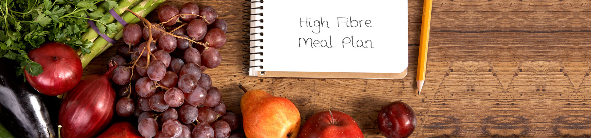 A wooden countertop with a selection of fruits and vegetables displayed alongside a notebook with the words "High Fibre Meal Plan".