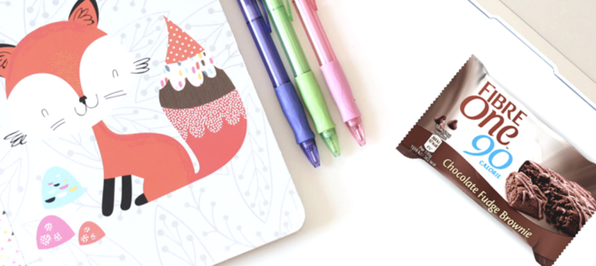 A table top with a notebook, a selection of pens and a Fibre One 90 Calorie chocolate fudge brownie.